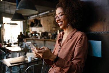 Kapalı kafede cep telefonu ve kulaklık kullanan afro saçlı genç siyahi bir kadın.