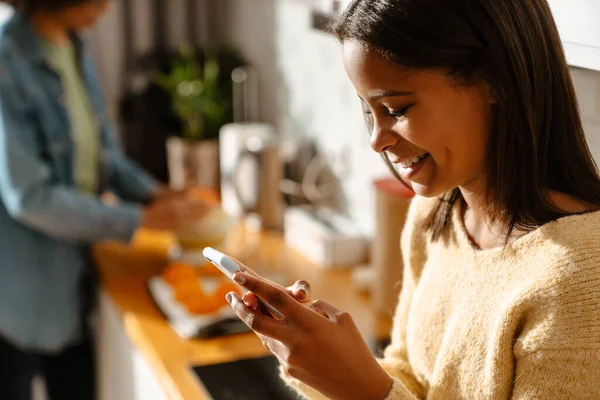 Stock image Black girl using mobile phone during spending time with her mother in kitchen at home