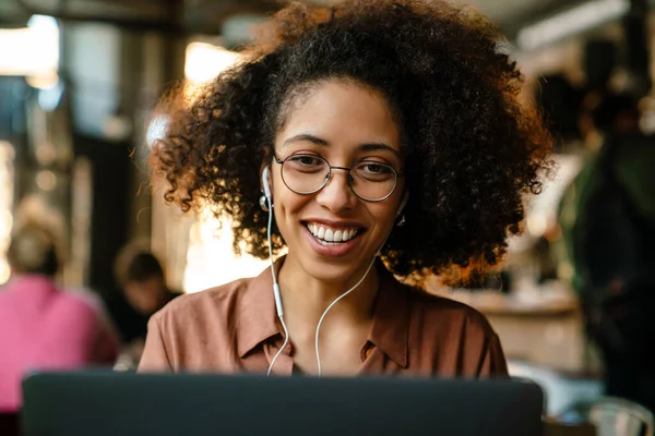 Mujer Afroamericana Joven Sonriendo Mientras Usa Portátil Los Auriculares Sentados —  Fotos de Stock