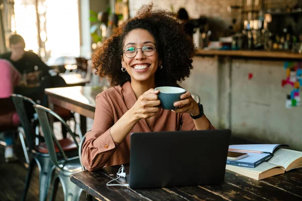 若いアフリカ系アメリカ人の女性がノートパソコンとコーヒーを飲みながらカフェに座っています — ストック写真