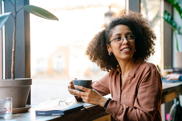 Young black woman with afro hairstyle smiling and drinking coffee in cafe indoors