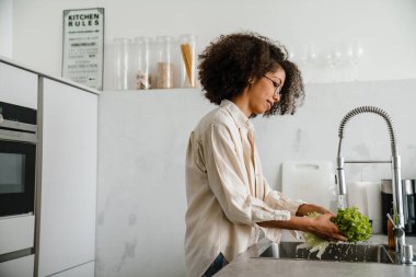 Siyah kadın gözlük takıyor, evde yemek yaparken marul yıkıyor.