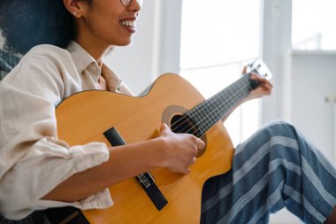 Evde oturmuş gitar çalan kıvırcık saçlı Afrikalı Amerikalı genç bir kadın.