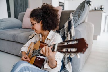 Evde oturmuş gitar çalan kıvırcık saçlı Afrikalı Amerikalı genç bir kadın.