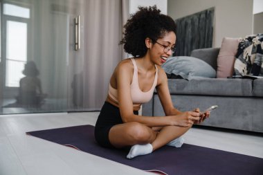 Evde yoga yaparken gözlük takan siyah bir kadın cep telefonu kullanıyor.