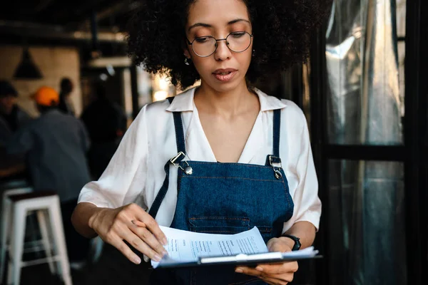 アフリカ系アメリカ人の若い女性が紙の書類を持ってクリップボードを持ちながらカフェで働く — ストック写真