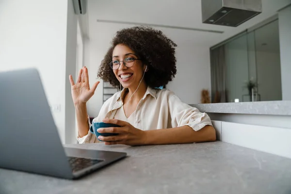 在家与笔记本电脑一起工作时 戴耳机喝咖啡的黑人妇女 — 图库照片