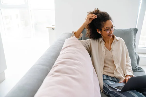 Wanita Hitam Dengan Earphone Bekerja Dengan Laptop Sambil Duduk Sofa — Stok Foto