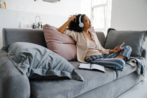 非洲裔美国女青年坐在沙发上 一边用手机和耳机 — 图库照片