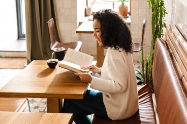 Genç siyah kadın kahve içiyor ve kafeteryada kitap okuyor.