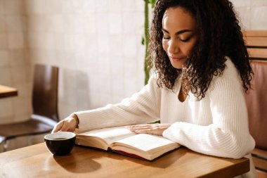 Genç siyah kadın kahve içiyor ve kafeteryada kitap okuyor.