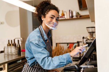 Yüz maskeli genç siyahi garson kapalı kafede kasiyerlik yapıyor.
