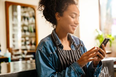 Kapalı kafede çalışırken cep telefonu kullanan genç zenci garson.