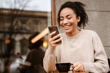 Açık kafede kahve içerken cep telefonu kullanan siyah bir kadın.