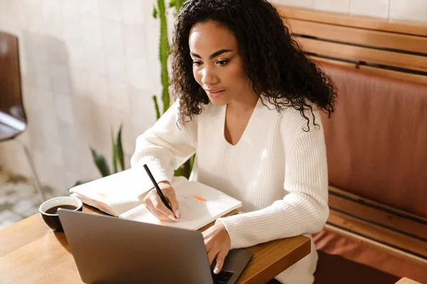 stock image Young black woman writing down notes while working with laptop in cafe indoors