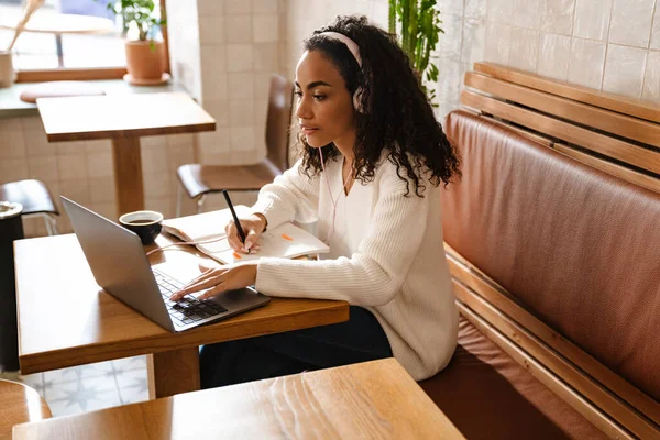 Young black woman writing down notes while working with laptop in cafe indoors