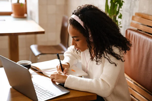 若い黒人女性書き留めノートをしながら作業ノートパソコンでカフェ室内 — ストック写真