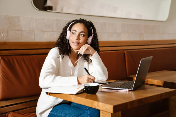 Young black woman writing down notes while working with laptop in cafe indoors