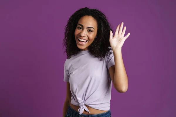 Tシャツを着た若い黒人女性が笑うと手を振って紫色の背景に孤立 — ストック写真
