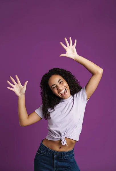 Young black woman with wavy hair laughing and dancing at camera isolated over purple background