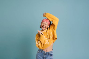 Kulaklıklı Asyalı kız mavi arka planda izole edilmiş cep telefonuyla müzik dinlerken dans ediyor.