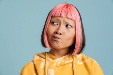 Pembe saçlı ve piercingli Asyalı kız mavi arka planda tek başına bakarken kaşlarını çatıyor.