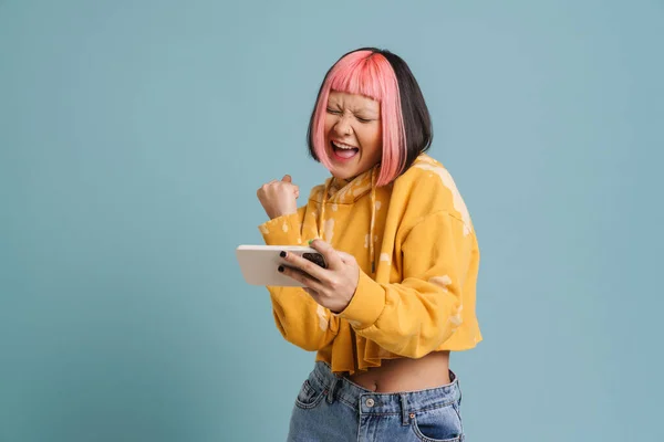 stock image Asian girl gesturing while playing online game on mobile phone isolated over blue background