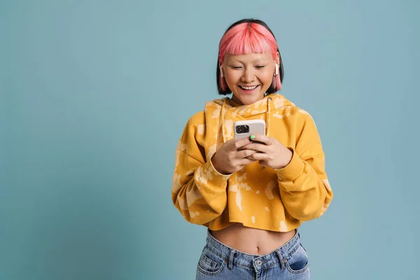 stock image Asian girl in earphones smiling and using mobile phone isolated over blue background