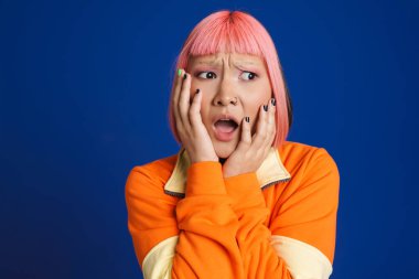 Pembe saçlı ve piercing takan Asyalı kız mavi arka planda izole edilmiş kameraya şaşırıyor.