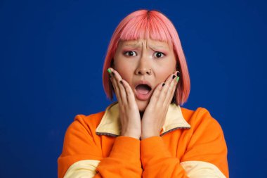 Pembe saçlı ve piercing takan Asyalı kız mavi arka planda izole edilmiş kameraya şaşırıyor.