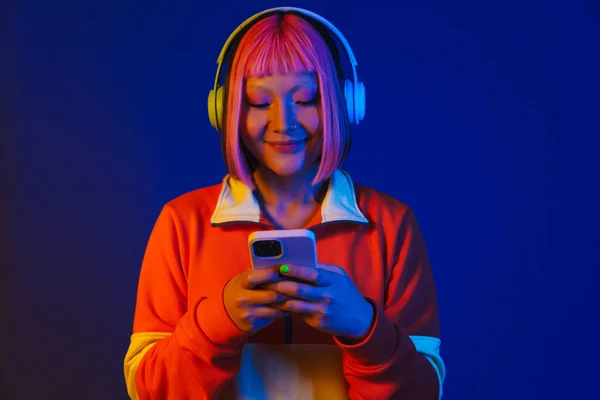 ヘッドフォンでアジアの女の子は笑顔と青の背景に隔離された携帯電話を使用して — ストック写真