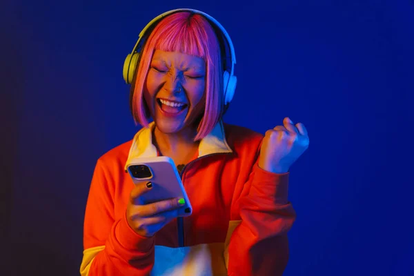 stock image Asian girl in headphones gesturing and using mobile phone isolated over blue background