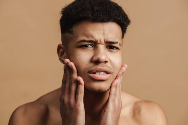 Yüzüne dokunurken kaşlarını çatan genç adam bej arka planda soyutlanmış.