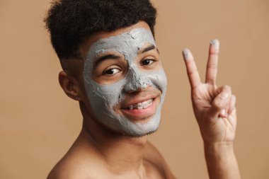 Kozmetik kil maskeli tişörtsüz adam bej arka planda izole edilmiş kameraya gülümsüyor ve el kol hareketi yapıyor.