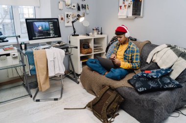 Kulaklıklı orta doğulu genç çocuk evdeki koltukta otururken dizüstü bilgisayar kullanıyor.