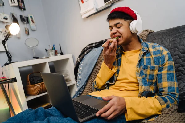 中东少年坐在家里沙发上 一边吃披萨 一边用笔记本电脑 — 图库照片