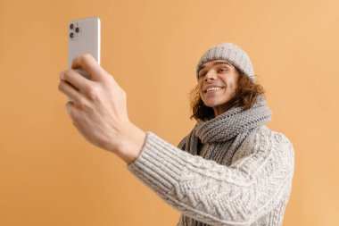Uzun saçlı, uzun saçlı, kışlık şapkalı ve eşarplı genç adam izole edilmiş kahverengi arka planda selfie çekiyor.