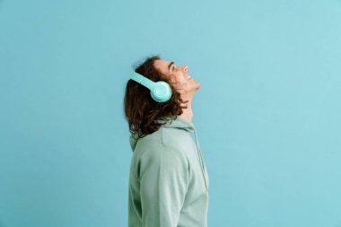 Uzun saçlı, kapüşonlu ve kulaklıklı genç yakışıklı adam mavi arka planda sağ tarafıyla ayakta duruyor.