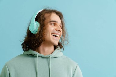 Kapüşonlu ve kulaklıklı genç yakışıklı adam kameraya bakıyor ve izole edilmiş mavi arka planda duruyor.