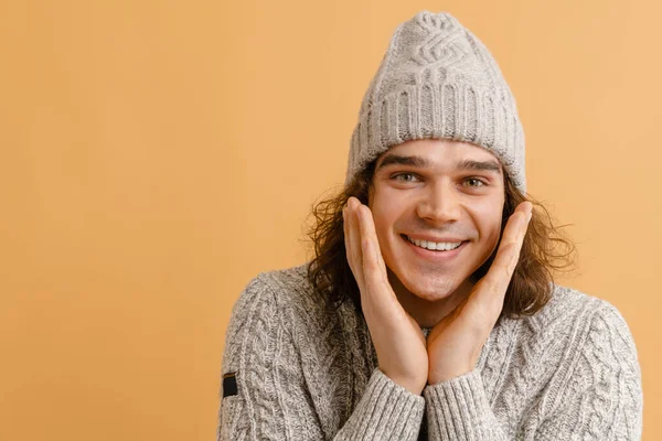 Młody Uśmiechnięty Mężczyzna Długimi Włosami Czapce Zimowej Trzymający Policzki Odizolowanym — Zdjęcie stockowe