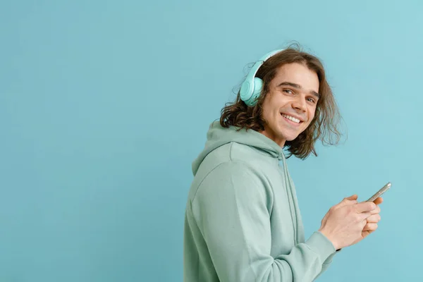 Młody Uśmiechnięty Mężczyzna Długimi Włosami Słuchawkach Trzymający Telefon Patrzący Kamerę — Zdjęcie stockowe