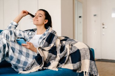 Evdeki kanepede dinlenirken cep telefonu kullanan genç bir kadın.