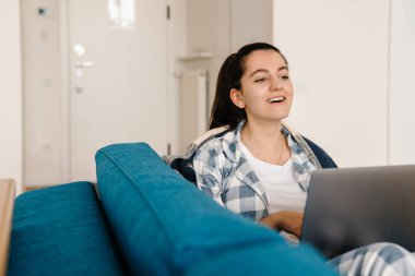 Evdeki kanepede dinlenirken gülümseyen ve laptop kullanan genç bir kadın.