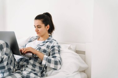 Evde yatağında dinlenirken pijama giyen genç bir kadın dizüstü bilgisayar kullanıyor.