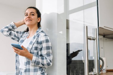 Evde dikilirken pijama giyen genç bir kadın cep telefonu kullanıyor.