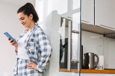 Evde dikilirken pijama giyen genç bir kadın cep telefonu kullanıyor.