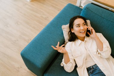 Genç, tatlı, gülümseyen Asyalı kız telefonda konuşuyor ve evdeki kanepede uzanıyor.