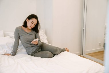 Evde takım elbiseli genç Asyalı kız telefonuyla evde yatağında oturuyor.