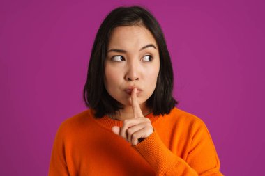 Asyalı genç bir kadın, mor arka planda izole edilmiş sessizlik jesti yaparken kaşlarını çatıyor.