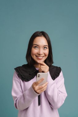 Gülümseyen ve cep telefonu kullanan Asyalı genç bir kadın mavi arka planda izole edilmiş.